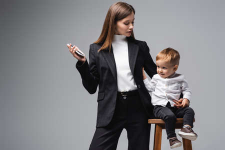 woman in black suit standing with smartphone near son sitting on high stool isolated on greyの写真素材