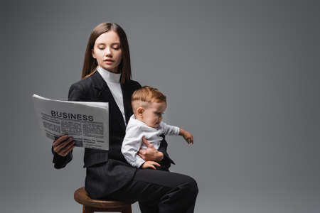 woman in black suit reading business newspaper while sitting with son on high stool isolated on greyの写真素材