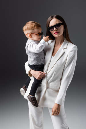trendy woman in sunglasses holding little son and looking at camera on greyの写真素材