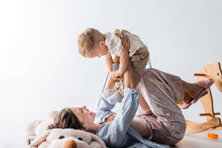 cheerful woman lying on toy dog and playing with toddler son on white