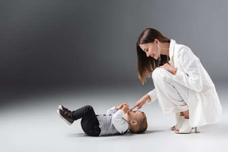 side view of cheerful woman in white suit touching nose of son lying on greyの写真素材