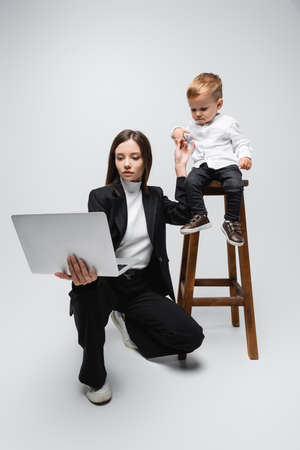 businesswoman with laptop holding hands with little son sitting on high stool on greyの写真素材