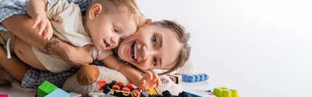 cheerful woman looking at camera while playing with son on white, banner