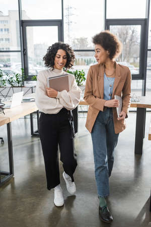 African american businesswoman holding laptop and talking to arabian colleague with documents in officeの写真素材