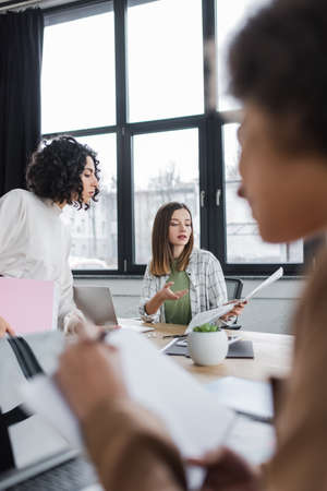 Businesswoman pointing at newspaper near laptop and muslim colleague in officeの写真素材