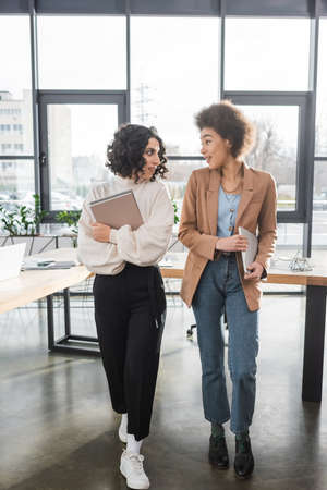 Positive interracial businesswomen with paper folders and laptop in officeの写真素材