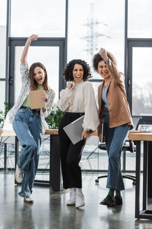 Excited multiethnic businesswomen with laptop and papers looking at camera in officeの写真素材