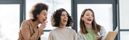Laughing multiethnic businesswomen looking at notebook in office, bannerの写真素材