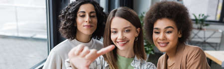Smiling businesswoman pointing with finger near multicultural colleagues in office, bannerの写真素材