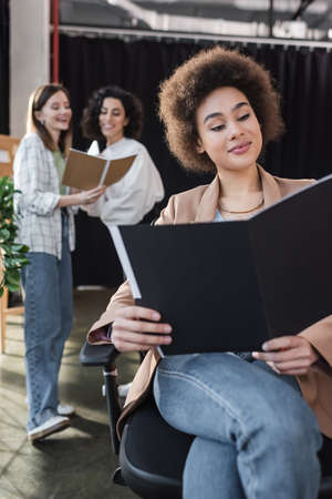 Cheerful african american businesswoman looking at paper folder near blurred interracial colleagues in officeの写真素材