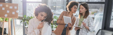 Arabian businesswoman talking on cellphone near multiethnic colleagues with digital tablet in office, bannerの写真素材