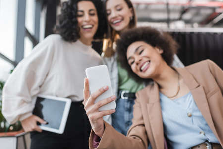 Blurred multiethnic businesswomen smiling while taking selfie on smartphone in officeの写真素材