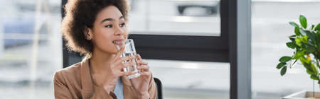 Positive african american businesswoman holding glass of water in office, bannerの写真素材