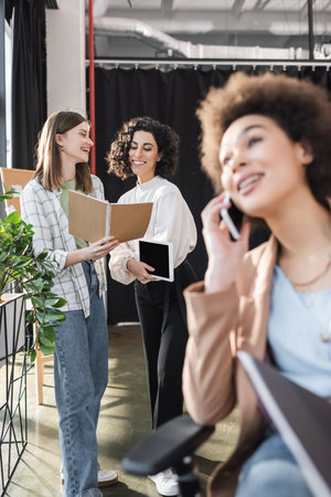 Positive multiethnic businesswomen looking at notebook while talking near blurred african american colleague talking on smartphone in officeの写真素材