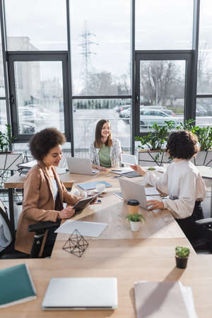 Positive interracial businesswomen talking near devices and papers on table in officeの写真素材