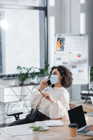 Arabian businesswoman in medical mask talking on smartphone in officeの写真素材