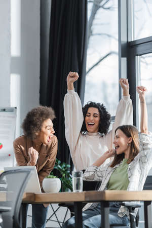 Excited multiethnic businesswomen showing yes gesture near laptop on table in officeの写真素材