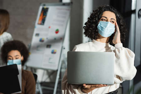 Muslim businesswoman in medical mask holding laptop while suffering from headache in office