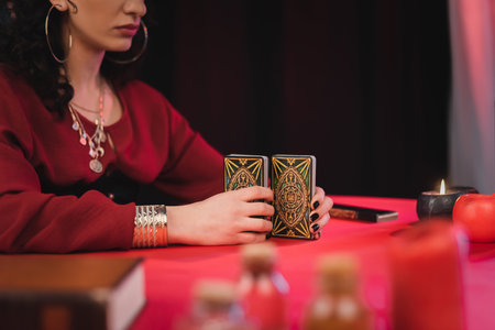 KYIV, UKRAINE - FEBRUARY 23, 2022: Cropped view of soothsayer holding tarot cards near blurred witchcraft supplies on table on black backgroundのeditorial素材
