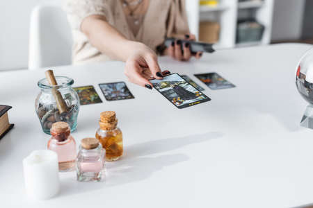 KYIV, UKRAINE - FEBRUARY 23, 2022: Cropped view of medium holding tarot card near jars and candle