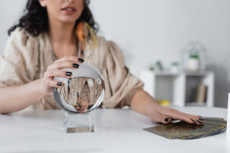 Cropped view of blurred fortune teller touching orb near tarot cards at home