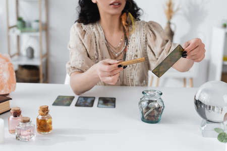 Cropped view of fortune teller holding sage stick and tarot card at home