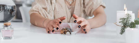 Cropped view of soothsayer holding magic crystals near candles and orb on table, banner