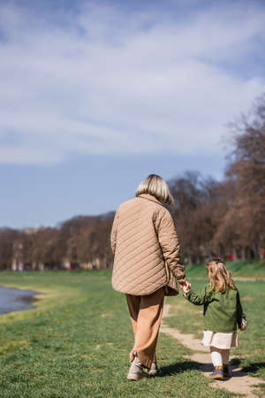 back view of mother and little daughter holding hands while walking in parkの写真素材