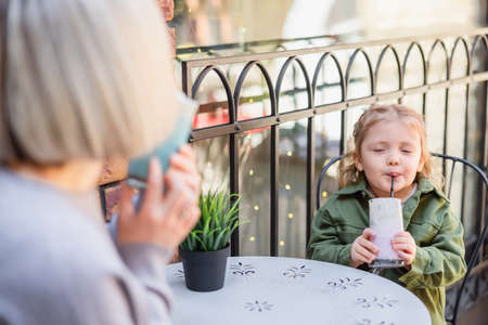 girl with closed eyes enjoying delicious dessert near blurred mom in cafe on streetの写真素材