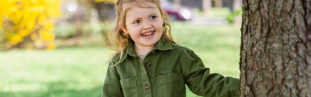 happy girl looking away while standing near tree in park, banner