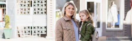 blonde woman in trendy outfit holding little girl outdoors, banner