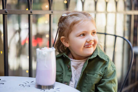 happy girl looking away near blurred glass of milkshake in street cafeの写真素材