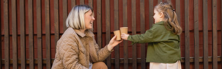 side view of cheerful woman and girl toasting with paper cups near wooden fence outdoors, banner