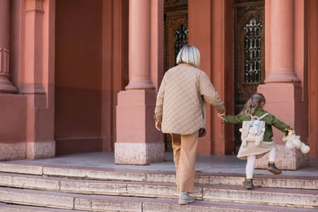 back view of woman and child with teddy bear holding hands while walking on stairs near buildingの写真素材
