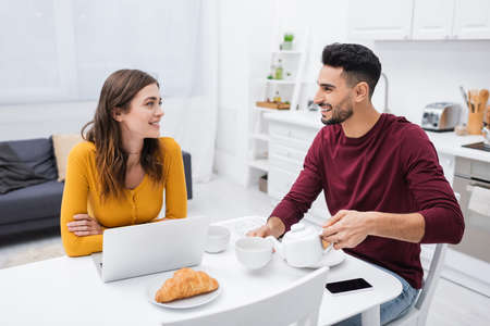 Happy multiethnic couple talking near devices and breakfast in kitchenの写真素材