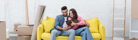 Cheerful interracial couple petting bengal cat on couch near packages at home, bannerの写真素材