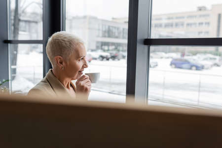 smiling businesswoman drinking coffee near office windows on blurred foreground
