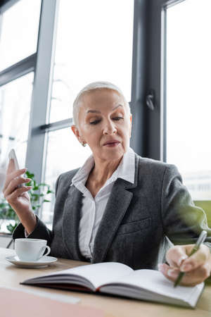 senior banker holding smartphone near coffee cup and writing in blurred notebookの写真素材