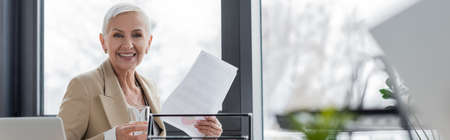 stylish banker with glass of water and document smiling at camera in office, bannerの写真素材