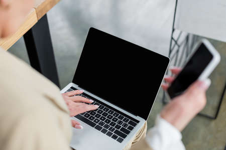 cropped view of banker with blurred smartphone typing on laptop with blank screen