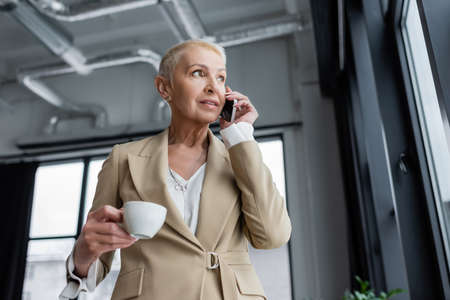 low angle view of stylish banker with coffee cup talking on smartphone in officeの写真素材