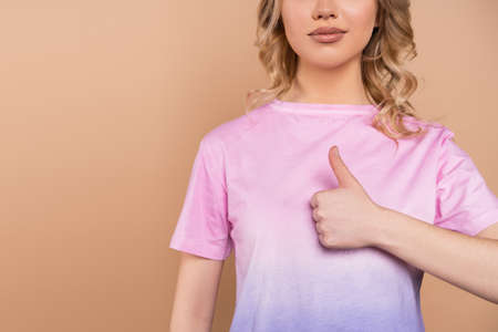 partial view of young woman in t-shirt showing thumb up isolated on beigeの写真素材