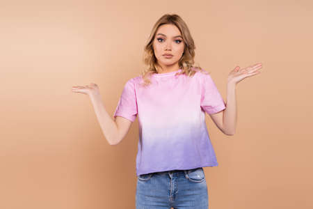 confused woman showing shrug gesture and looking at camera isolated on beige