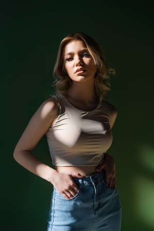 sensual woman in beige top standing with hands in pockets of jeans on dark background