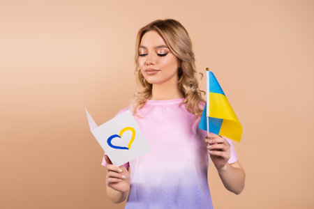 woman reading card with blue and yellow heart and holding small ukrainian flag isolated on beigeの写真素材