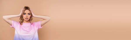 upset woman holding hands on head while looking at camera isolated on beige, bannerの写真素材