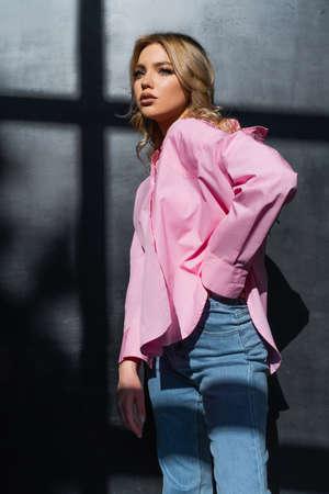 stylish woman in pink shirt standing with hand in pocket of jeans near black wall with shadowsの写真素材