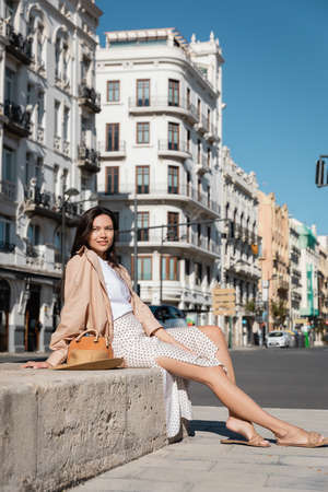 full length of brunette woman in skirt sitting on city street near handbag and straw hatの写真素材