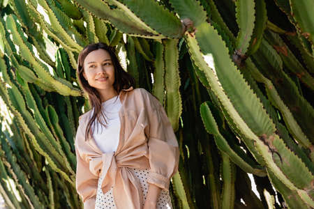 low angle view of brunette woman in shirt smiling near giant cactiの写真素材