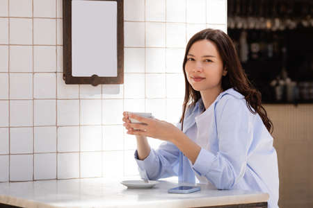 brunette woman with cup of coffee looking at camera on cafe terrace